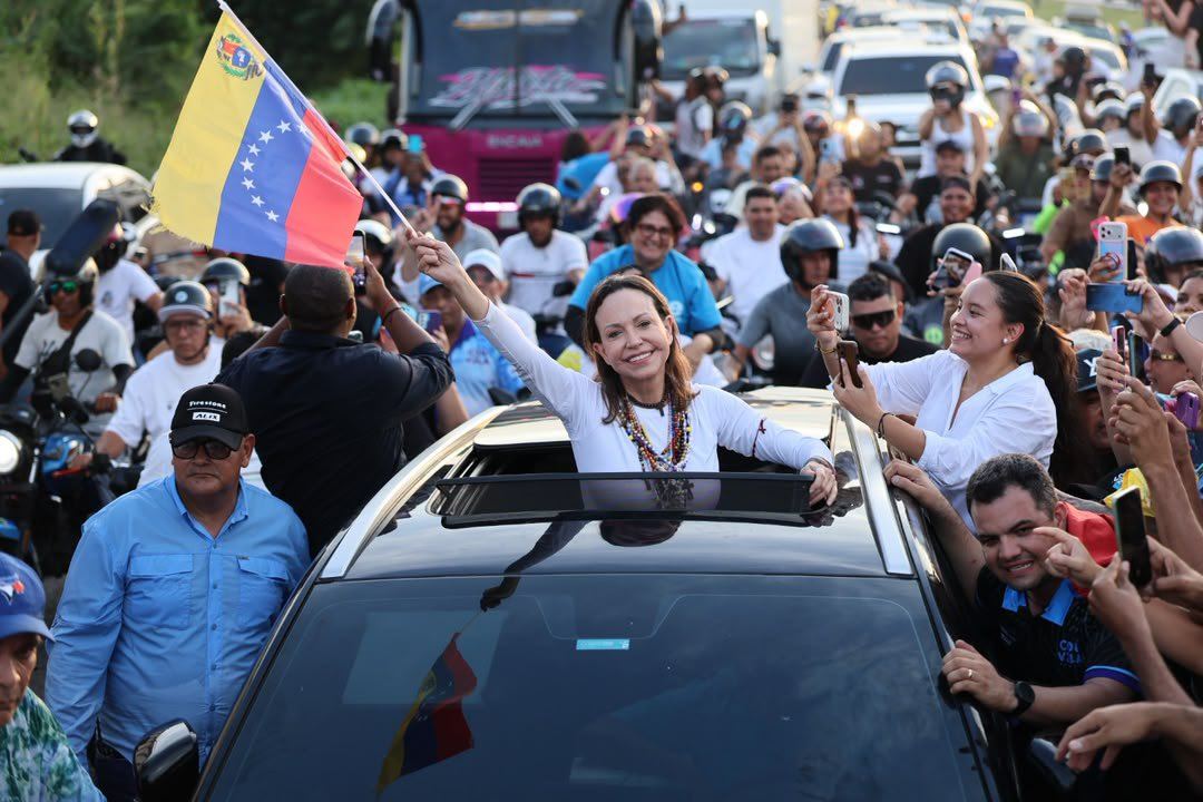 María Corina Machado saluda desde un vehículo a seguidores que llevan banderas de Venezuela
